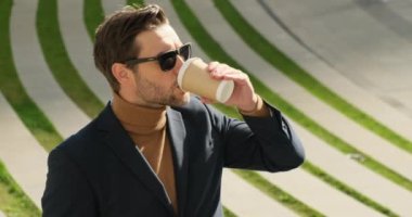 Yakışıklı, takım elbiseli, sokakta elinde kahveyle dolaşan. Takım elbiseli şık bir işadamı ve boğazlı kazak elinde kahve ve tatilin tadını çıkarıyor. Güzel saç stili ve güneş gözlüklü yakışıklı adam.