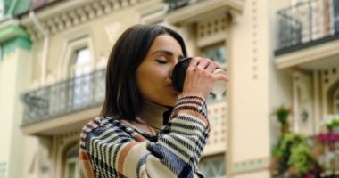 Uzun kıvırcık saçlı, cappuccino içen güzel, mutlu bir kadın. Sokakta gün ışığında sabah kahvesi içen neşeli bir kadın. Gri elbiseli şık bir kadın Paris 'te açık havada kahve içiyor.