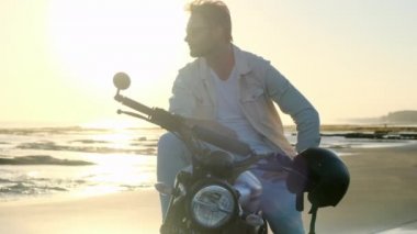 Okyanusta motosiklet süren şık bir adam. Vahşi bir şehir yaşam tarzı. Siyah motosikletli sakallı ve bıyıklı adam güneş gözlüklerini gün batımında klasik kafe yarış motosikletinde çıkardı..