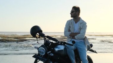 Okyanusta motosiklet süren şık bir adam. Vahşi bir şehir yaşam tarzı. Siyah motosikletli sakallı ve bıyıklı adam güneş gözlüklerini gün batımında klasik kafe yarış motosikletinde çıkardı..