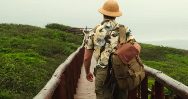 Sırtında sırt çantasıyla gezgin bir erkek tropikal otların arasında ahşap bir köprü boyunca yürüyor. Safari tarzı, şapkalı ve gömlekli bir adam Amazon 'un ünlü yerlerine seyahat ediyor. seyahat blogcusu tatilde 