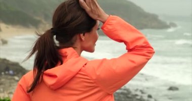 Yoğun fitness antrenmanından sonra soluklanıyor. Sıcak Işık. Spor ceketli bir kız okyanusta duruyor ve büyük dalgalara bakıyor. Gezgin kız dağın zirvesinde duruyor.