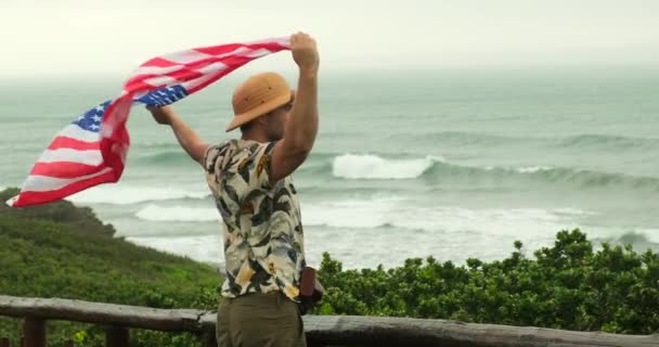 崖の端にある冒険的な男は 夕日の間にキャニオンの美しい景色を見ています アメリカンフラッグを誇るサファリスタイルのスタイリッシュな旅行者が海を楽しむ — ストック動画