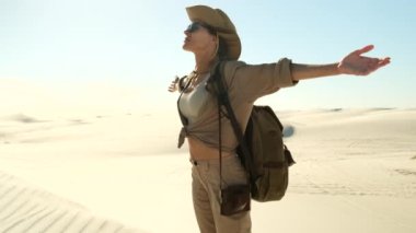 Sunset over the sand dunes in the desert. a girl tourist with a travel backpack spread her arms to the sides, feeling a sandstorm in the desert. woman in safari clothes enjoying a trip to africa