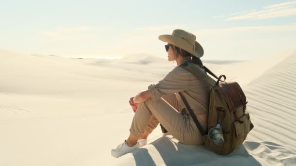 サファリの帽子とサングラスの美しい旅行者は暑い太陽の下で砂漠を遠く見ています 澄んだ青空とオレンジの砂丘砂漠の風景 砂漠の真ん中で一人旅をしていると — ストック動画