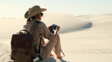 woman in travel clothes sits on sand dune in the desert and takes a photo on a film camera. travel female enjoys traveling alone through the desert in africa. girl in boots and a hat sits on the sand