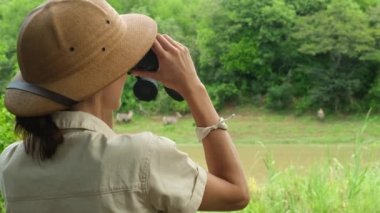 young people watch and photograph wild elephants on a safari tour in a national park. rear view tourist girl looking through binoculars at tropical animals in the African savannah. girl in safari hat