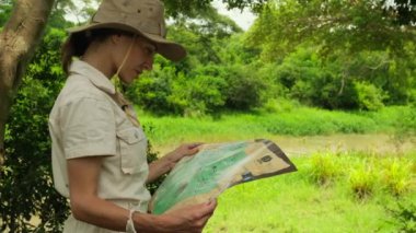 a girl tourist in travel overalls and a safari hat stands near the Amazon and looks at the route on the map. a traveler in the savannah in africa explores routes among the jungle in the national park