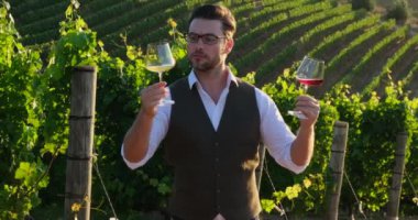 millennial vintner holding a glass and a bottle of organic bio red wine outdoors in a vineyard. stylish man in a vest and shirt stands on a grape plantation and holds two glasses of white and red wine