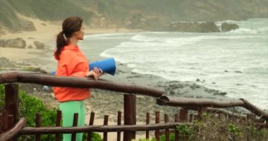 Dişi, gücünü ve ruhunu geri kazanmak için spor egzersizleri yapıyor. Yoga pozisyonu. Spor ceketli kız yoga minderi tutuyor ve taze deniz esintisinin tadını çıkarıyor. Genç ve güzel bir kadın sahilde meditasyon yapıyor.