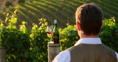 wine bottle and glass on wodden barrel. on a wooden table there is a bottle of wine and a glass of red wine at sunset, a man shows a heart of hands. sommelier waiting for his new vintage of grapes