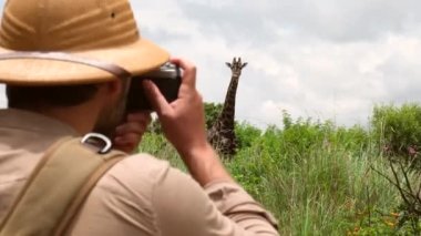 man traveler in safari style photographs a giraffe in the savannah. Happy zoology student men taking photo while giraffe drinking from lake. traveler guy watching giraffes in the savannah