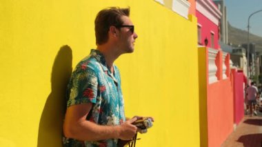 tourist man smiling happy using vintage camera at the city. man taking photographs on SLR. stylish man in a summer shirt makes a photo against the background of colored houses and smiles at the camera