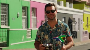 tourist man smiling happy using vintage camera at the city. man taking photographs on SLR. stylish man in a summer shirt makes a photo against the background of colored houses and smiles at the camera