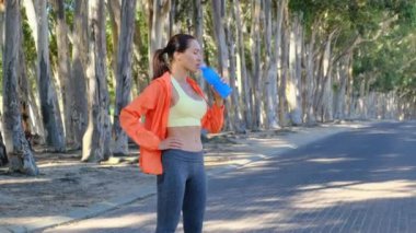 beautiful girl drinking water. Fitness athlete young american woman listening to music on earphones drinking water in a reusable water bottle after working out exercising on sunny day at the park