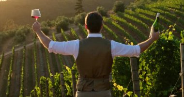 Portrait of young, millennial vintner holding and smelling a glass of organic bio red wine outdoors in a vineyard. male sommelier in a white shirt and vest holding a bottle of wine and a glass of wine