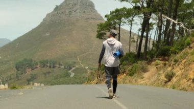 a sporty man in shorts and sports leggings walks along a road in a park near high mountains and holds a yoga mat and a jump rope in his hands. stylish sportsman man walking after jogging with yoga mat