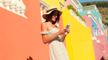 People lifestyle concept. Using mobile phone, typing sms message. a beautiful girl in a summer dress stands near colorful houses in cape town south africa and texts her boyfriend