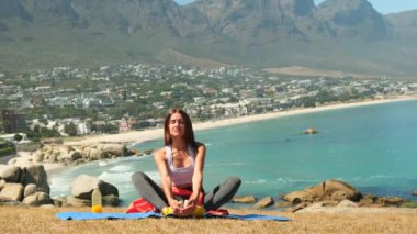 sportswoman doing exercises and doing yoga fitness, sport and healthy lifestyle. athletic woman in leggings sits in a lotus position on a yoga mat on green grass, mountains and the ocean in cape town