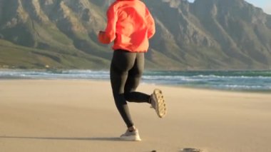 Koşucu güneşli parlak ışıkta koşuyor. Dışarıda kadın fitness eğitimi var. Güzel bir spor kızı okyanus ve dağların yakınındaki vahşi bir plajda koşuyor. Kumsalda kumda koşan Yatletik kız.