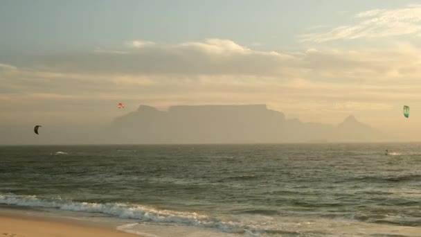日の出のビーチとテーブル山の風景 南アフリカのテーブルマウンテンを背景に サーフィンボード上のプロのウィンドサーファーのセーリング人の会社はカイトサーフィンに乗る — ストック動画