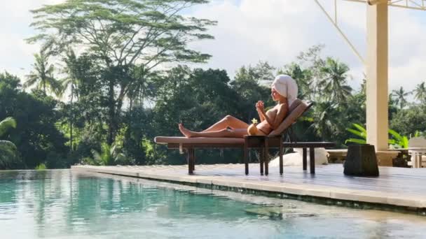 頭にタオルを着た女の子が休暇中にプールの近くのサンラウンジャーでマニキュアをしています プールの近くの太陽の下で日光浴をして爪のファイルでマニキュアをしてる — ストック動画