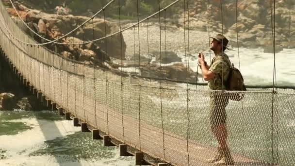 ティツィカメマ国立公園のルートで嵐の川の口の吊り橋 男性写真家が手にカメラを持ち南アフリカの吊り橋を見て — ストック動画