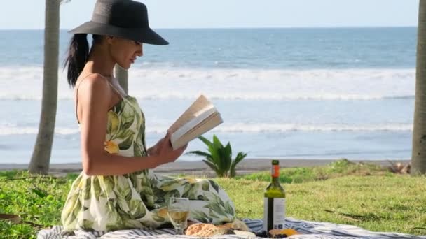 帽子とドレスの美しい女の子が草の上に座って本を読んでいます ピクニックの時間だ 公園でピクニックを楽しんでる女の子 バリで休暇中の女性がワインを片手にピクニックをしました — ストック動画