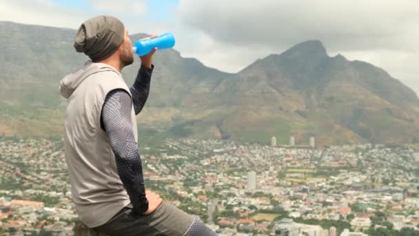 屋外のスタジアムでウォーターボトルを持ってるアメリカ人男性 帽子とTシャツの運動選手はジョギングの後 山の公園のボトルから水を飲みます 筋肉の男はトレーニングから休憩を取って — ストック動画