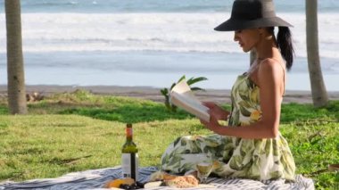 Şapkalı ve elbiseli güzel bir kız çimlere oturur ve kitap okur. Piknik zamanı. Parkta piknik yapan bir kız. Bali 'de tatil yapan bir kadın bir bardak şarapla piknik yapmış.