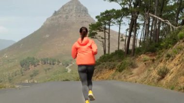 Şehir sokaklarında koşan genç bir kadın. Spor kıyafetleri içinde atletik bir vücudu olan atletik bir kadın sabahın erken saatlerinde parktaki yolda dağların arka planına doğru koşar.