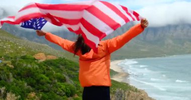 Amerikan bayrağını denizin önünde tutan genç bir kadının arkadan görünüşü, bağımsızlık günü konsepti. Amerikan bayrağı taşıyan çekici bir kadın eğleniyor ve gününü geçiriyor. Okyanusta gezgin bir spor kızı.