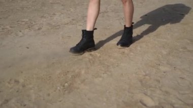 Siyah deri ayakkabılı bir kız kumsalda yürüyor ve kumu tekmeliyor..