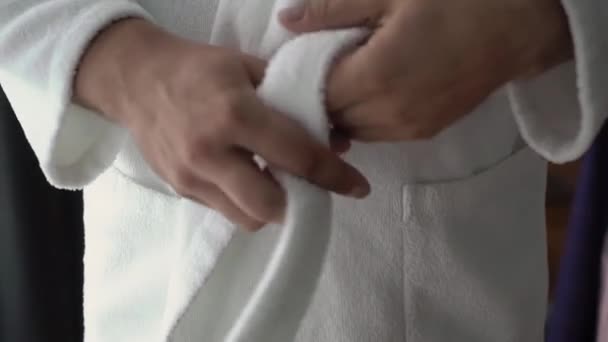The man puts on a white terry robe, ties a belt. Morning after waking up. – stockvideo
