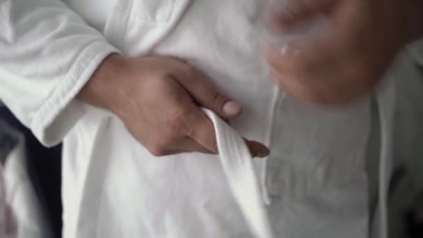 The man puts on a white terry robe, ties a belt. Morning after waking up. — Stockvideo