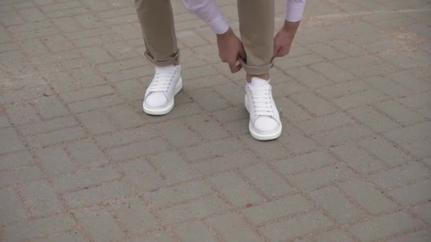 A young man in white sneakers and beige trousers is turning up his trousers. — Video