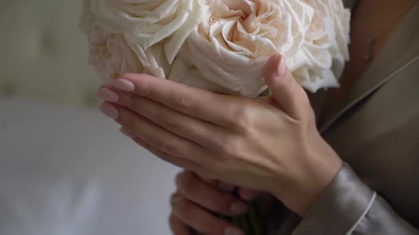 A bride in a gray robe holds a bouquet of white rose flowers in her hands — ストック動画