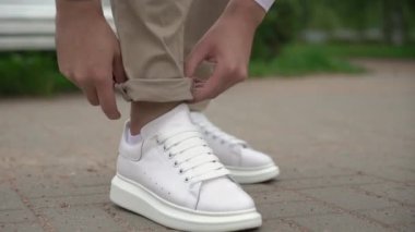 A young man in white sneakers and beige trousers is turning up his trousers.