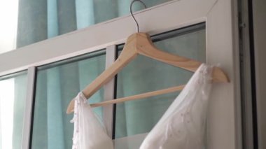 Wedding dress hangs on a hanger in the bedroom by the window. Bride takes gown.