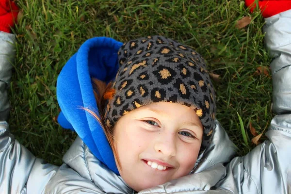 푸른 봄 초원의 잔디 위에 누워 있는 행복 한 어린 소녀, 귀여운 소녀. 아이는 표범 모양의 모자, 은색 자켓, 파란색 재킷, 빨간 장갑을 착용하고 있다. 즐거운 행복 한 아이 — 스톡 사진