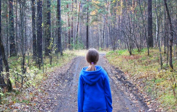 En flicka går ensam när hon går genom skogen en höstdag. Ensamhet och melankoli. En ung flicka i blå tröja med huva går längs en stig bland träden i skogen med sin — Stockfoto
