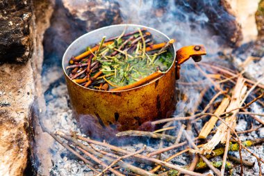 Vahşi doğada hayatta kalmak. Kamp ateşi arka planı. Ateşin üstünde vişneli çay çubuklarıyla tava. Vahşi doğada çay pişirmek....
