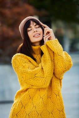 Dışarıda sarı yün kazak ve şapka giymiş güzel bir Avrupalı esmer. Kalın ve şık sonbahar kıyafetleri giyen güzel kız serin havada..