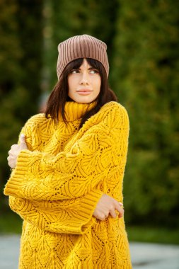 Dışarıda sarı yün kazak ve şapka giymiş güzel bir Avrupalı esmer. Kalın ve şık sonbahar kıyafetleri giyen güzel kız serin havada..