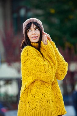 Dışarıda sarı yün kazak ve şapka giymiş güzel bir Avrupalı esmer. Kalın ve şık sonbahar kıyafetleri giyen güzel kız serin havada..