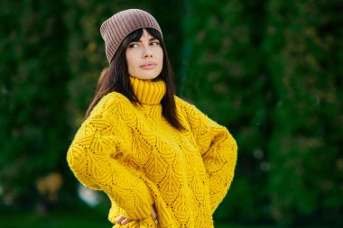 Dışarıda sarı yün kazak ve şapka giymiş güzel bir Avrupalı esmer. Kalın ve şık sonbahar kıyafetleri giyen güzel kız serin havada..