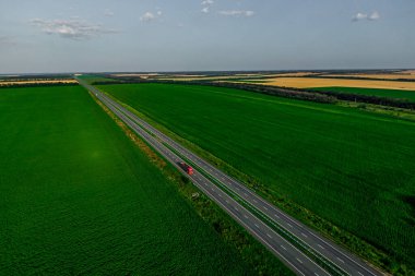 Yalnız kırmızı kamyon asfalt yolda gün batımında yeşil tarlalar boyunca sürüyor. Havadan görüldü. Hava manzaralı. İHA fotoğrafçılığı. kargo teslim