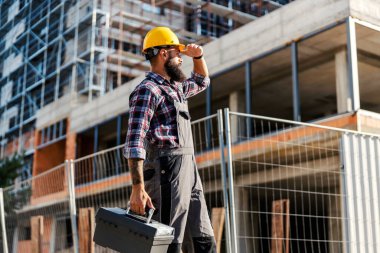 A hardworking worker with a toolbox in hand is arriving at a construction site. He is looking at the site and, he is ready to do his job. clipart