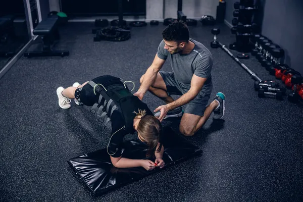 EMS training studio. Shot from above of a female athlete wearing an EMS suit and a male trainer in grey sportswear helping and showing her the exercise for plank position Electrical muscle stimulation