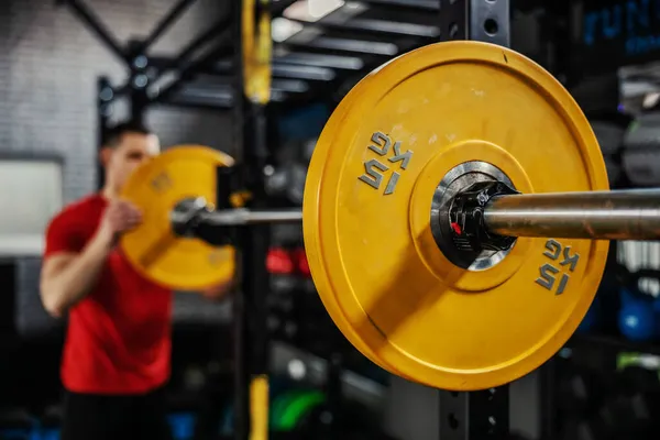 Preparing barbell weight for strong muscle burning training. A man in red sportswear sets up barbell weights in the gym. Close-up shot of a yellow barbell on a barbell. Blurred background